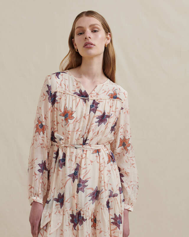 studio photo of a model wearing a long sleeve maxi dress by fashion brand Analia. The dress is a relaxed fit, full length button front, gathered drop waist flowing skirt, round neckline, chest piping detail with delicate gatherings and a self-fabric sash to cinch the waist. Cream viscose fabric with a purple, orange, blue bloom print.