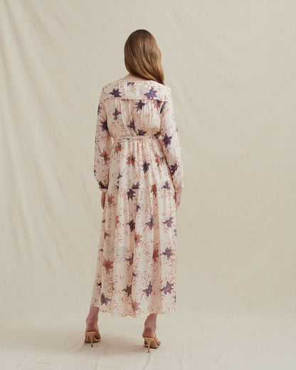 studio photo of a model wearing a long sleeve maxi dress by fashion brand Analia. The dress is a relaxed fit, full length button front, gathered drop waist flowing skirt, round neckline, chest piping detail with delicate gatherings and a self-fabric sash to cinch the waist. Cream viscose fabric with a purple, orange, blue bloom print