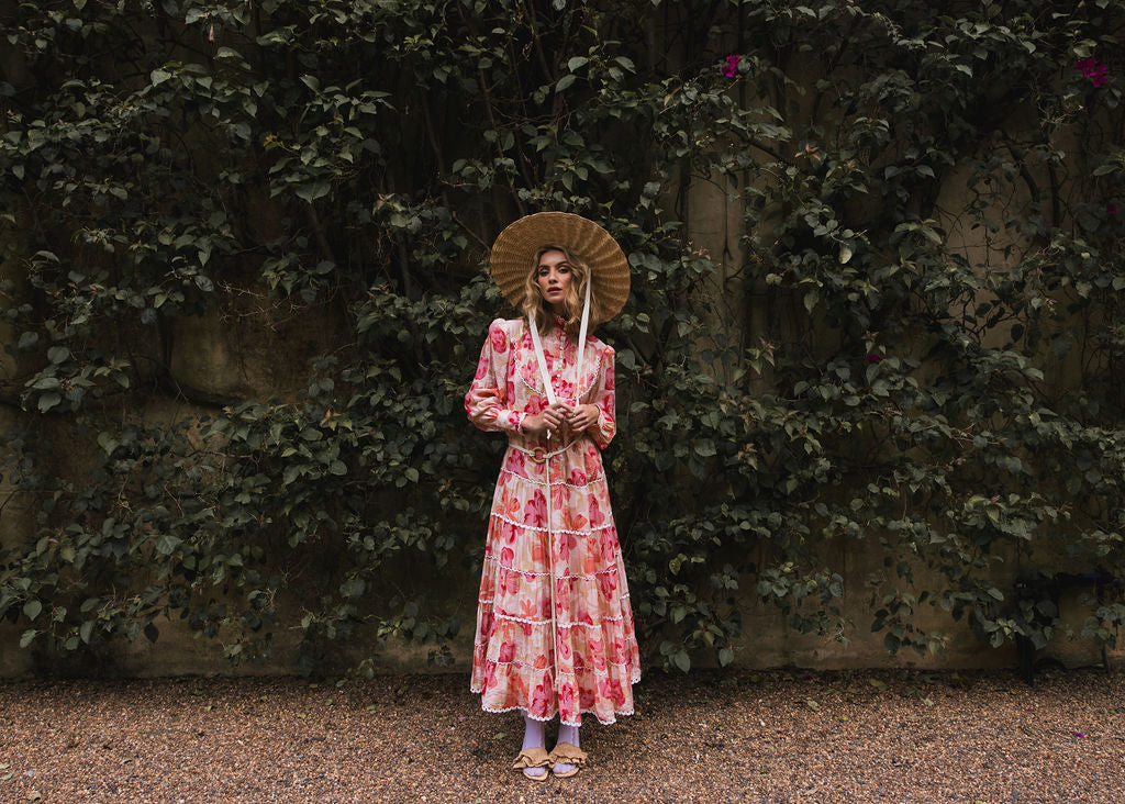 Campaign image by Analia the label, collection two, Tuscan Bloom. The beautiful model is wearing the Charlotte maxi dress, Summer Meadow. The dress has a gathered tiered maxi skirt, pleated bodice, blouson cuffed sleeves, stand collar and shell button front from the dropwaist, in a soft peach and pink floral print on a cream lightweight viscose fabric and completed with white cotton lace trim.