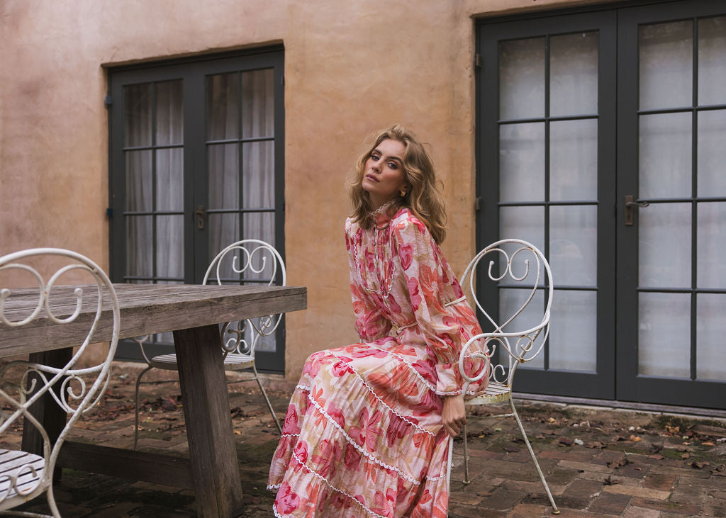 A elegant woman sitting in a Tuscan Bloom setting wearing the Charlotte Maxi Dress Summer Meadow by Analia the label. Maxi length with a gathered tiered skirt, blouson sleeve and cuff, pleated bodice, stand collar in a lightweight viscos fabric with a light peach and pink floral pattern on a cream base, beautifully completed with white cotton lace. 