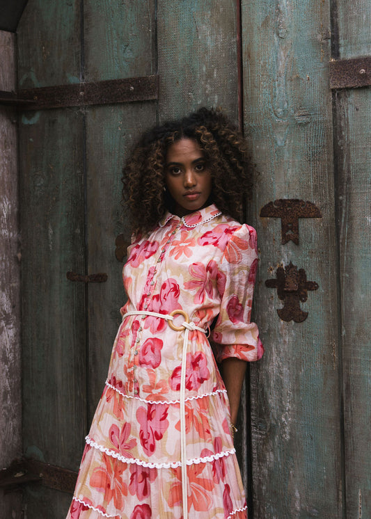 Beautiful model wearing a blouson sleeve maxi dress with full gathered tiers with lace detail, pea collar with lace detail, front shell buttons to the drop waist with a natural rope and round wood buckle in an organic lightweight viscose on a print of blooms in soft cream, peach and pink.